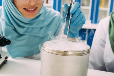 partial vie of female muslim scientist holding tweezers during experiment with dry ice in laboratory clipart