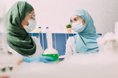 female muslim scientists with flask looking at each other while experimenting with dry ice in chemical laboratory clipart