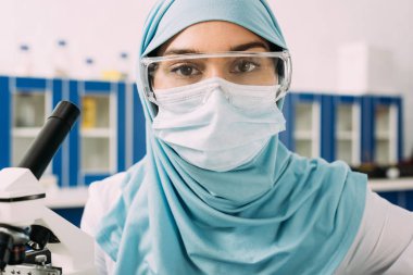 tıbbi maske, gözlük ve laboratuvar kamerada bakarak Türban kadın Müslüman bilim adamı