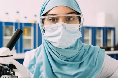 female muslim scientist in medical mask, goggles and hijab looking at camera in laboratory clipart