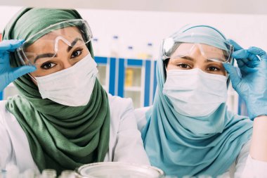 female muslim chemists in medical masks and hijab looking at camera in laboratory clipart