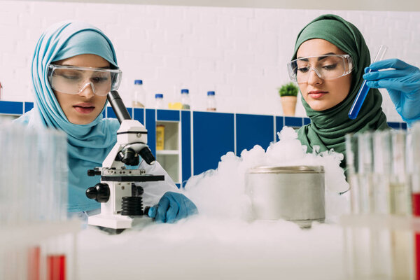 женщины-ученые-мусульмане экспериментируют с микроскопом, пробиркой и сухим льдом в химической лаборатории
