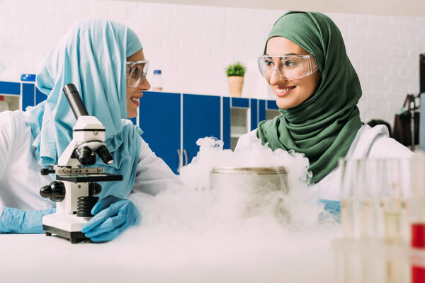 smiling female muslim scientists experimenting with microscope and dry ice in chemical laboratory