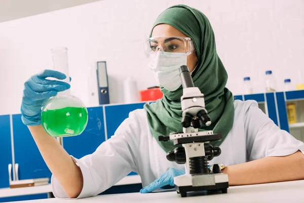 Cientista Muçulmano Fêmea Sentada Mesa Com Microscópio Segurando Frasco Durante — Fotografia de Stock