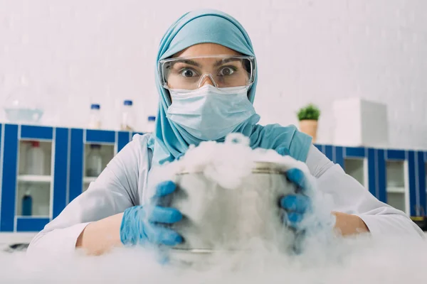 研究室でドライアイスでポットを押しながらカメラを見て驚く女性イスラム教徒の科学 — ストック写真