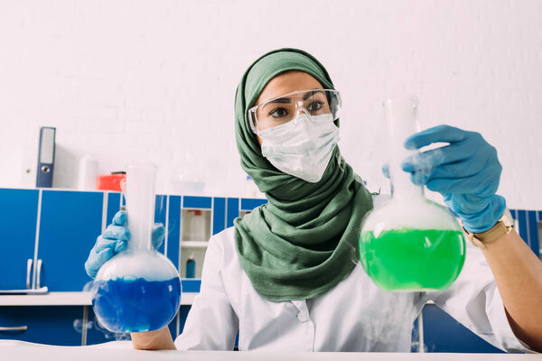 female muslim scientist in medical mask holding flasks during experiment in chemical laboratory