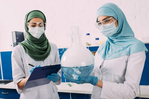 Científicos Musulmanes Femeninos Concentrados Máscaras Médicas Que Sostienen Portapapeles Líquido —  Fotos de Stock