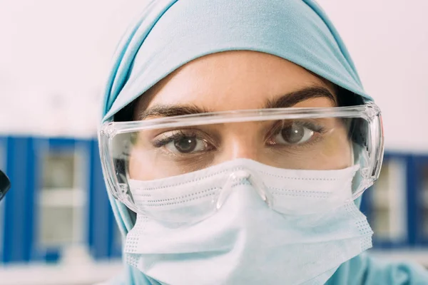 关闭女性穆斯林科学家在医疗面具 护目镜和头巾看着相机 — 图库照片