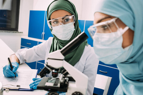 female muslim scientists using microscope and writing in clipboard during experiment in chemical laboratory