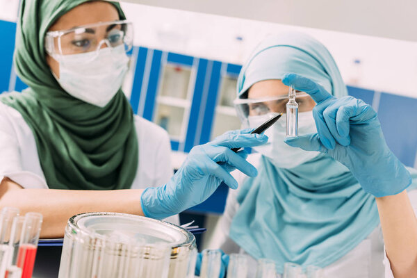 female muslim scientists in medical masks holding glass ampoule during experiment in chemical laboratory