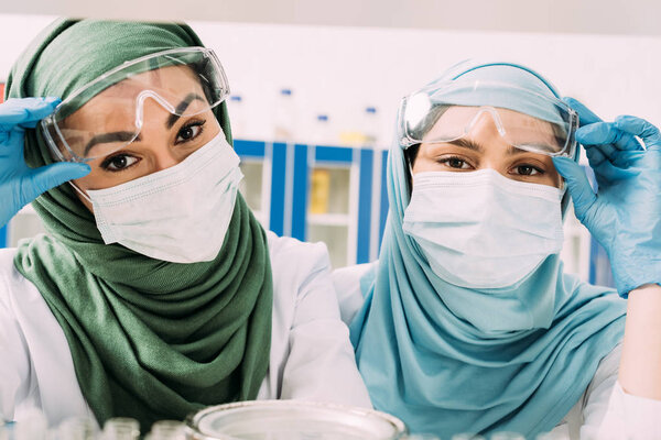 female muslim chemists in medical masks and hijab looking at camera in laboratory