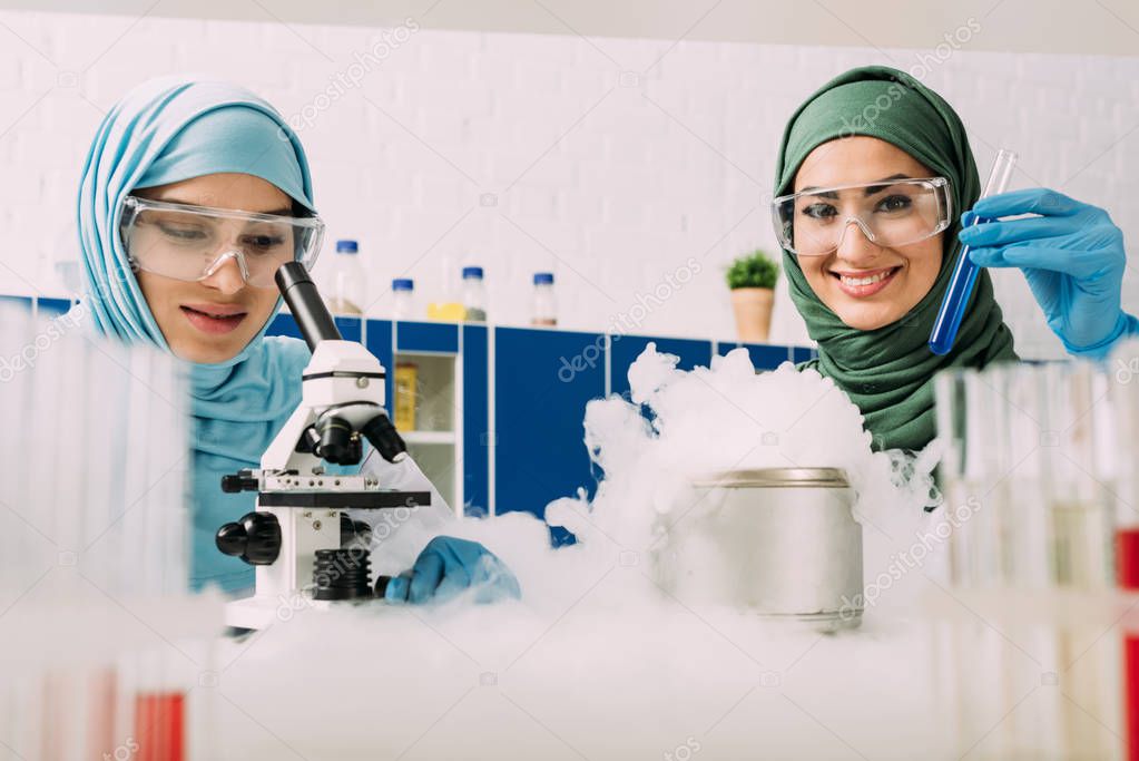 female muslim scientists in goggles experimenting with microscope and dry ice in chemical laboratory