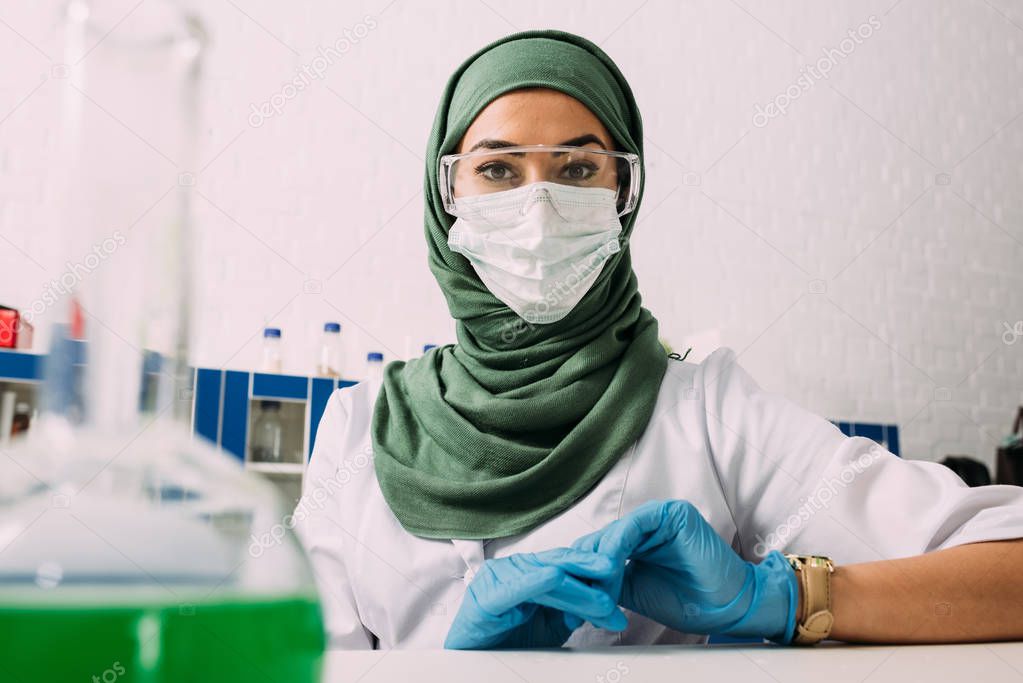 female muslim scientist in goggles and medical mask looking at camera in chemical laboratory with flask on foreground 