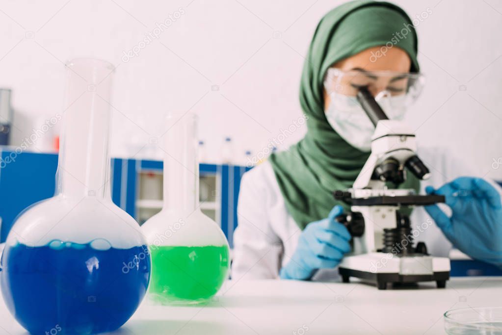 female muslim scientist sitting at table with flasks and using microscope during experiment in chemical laboratory