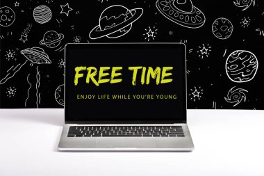 laptop on table with enjoy life while you are young and free time lettering on screen with white galaxy illustration on black  clipart
