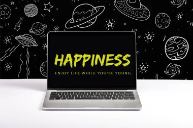 laptop on table with enjoy life while you are young and happiness lettering on screen with white galaxy illustration on black  clipart