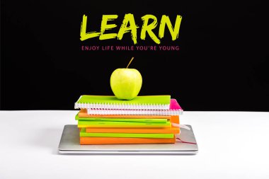 closed laptop, pile of notebooks with green apple on top with learn and enjoy life while you are young lettering on black  clipart