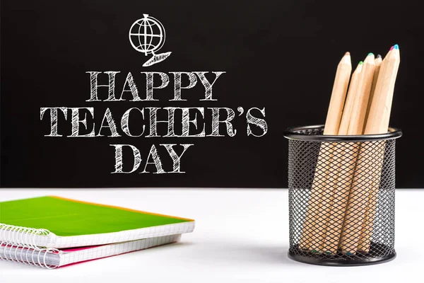 笔记本和彩色铅笔在桌子上与白色快乐的老师天在黑色上的字母 — 图库照片