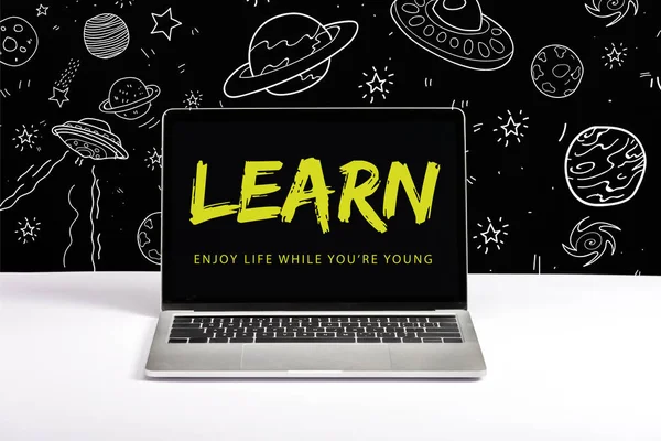 laptop on table with enjoy life while you are young and learn lettering on screen with white space illustration on black 