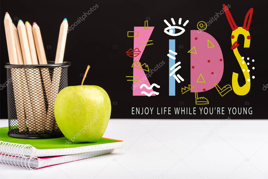 green apple, notebooks and color pencils on table with enjoy life while you are young and kids lettering on black 