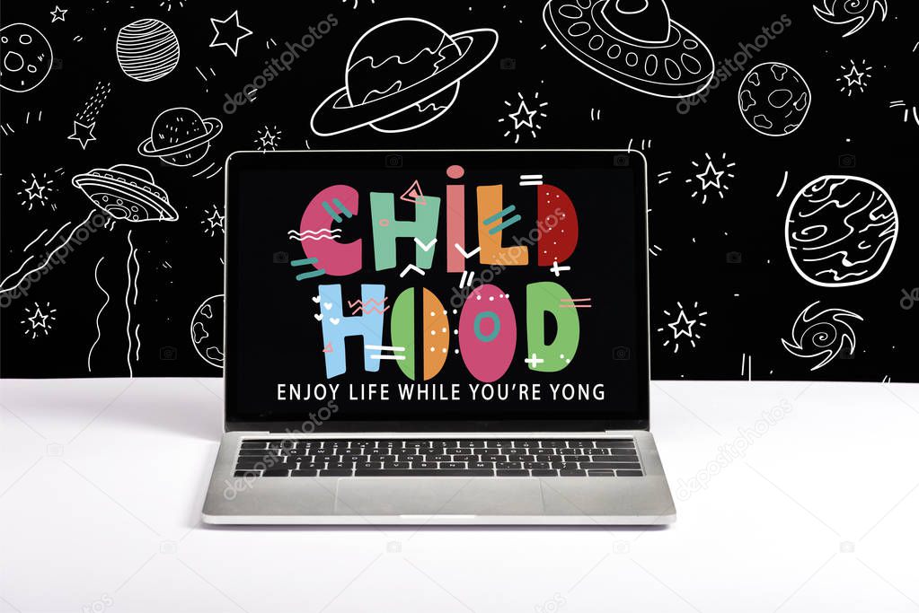 laptop on table with enjoy life while you are young and childhood lettering on screen with white space illustration on black 