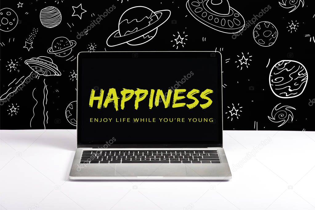laptop on table with enjoy life while you are young and happiness lettering on screen with white galaxy illustration on black 
