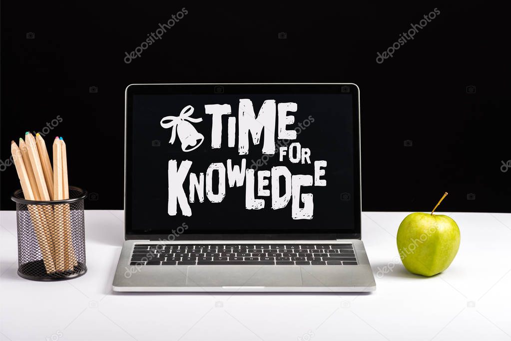 fresh green apple, color pencils and laptop on table with time for knowledge lettering and bell on screen isolated on black