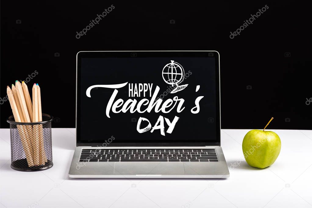 fresh green apple, color pencils and laptop on table with happy teachers day lettering and globe on screen isolated on black