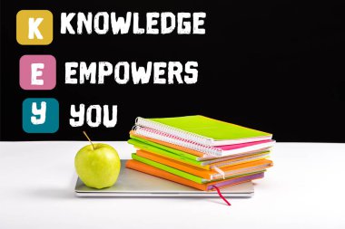 closed laptop, green apple, notebooks and color pencils on desk with knowledge empowers you lettering on black  clipart