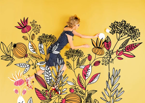 花のイラストと黄色の背景に横たわっているソーサーとカップを持つ若いエレガントな女性のトップビュー — ストック写真