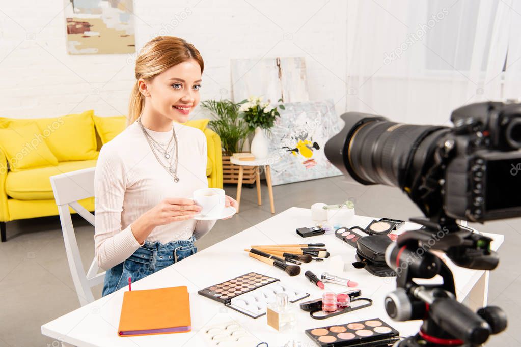 beauty blogger sitting at table with decorative cosmetics and holding cup of coffee in front of video camera