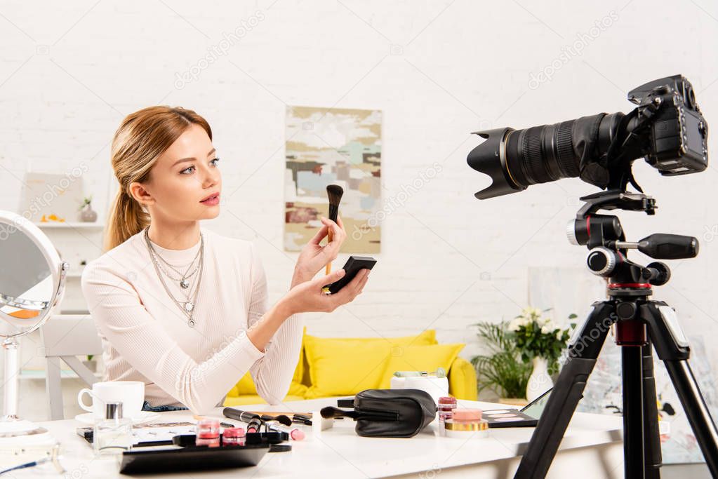 beauty blogger holding face powder and cosmetic brush in front of video camera