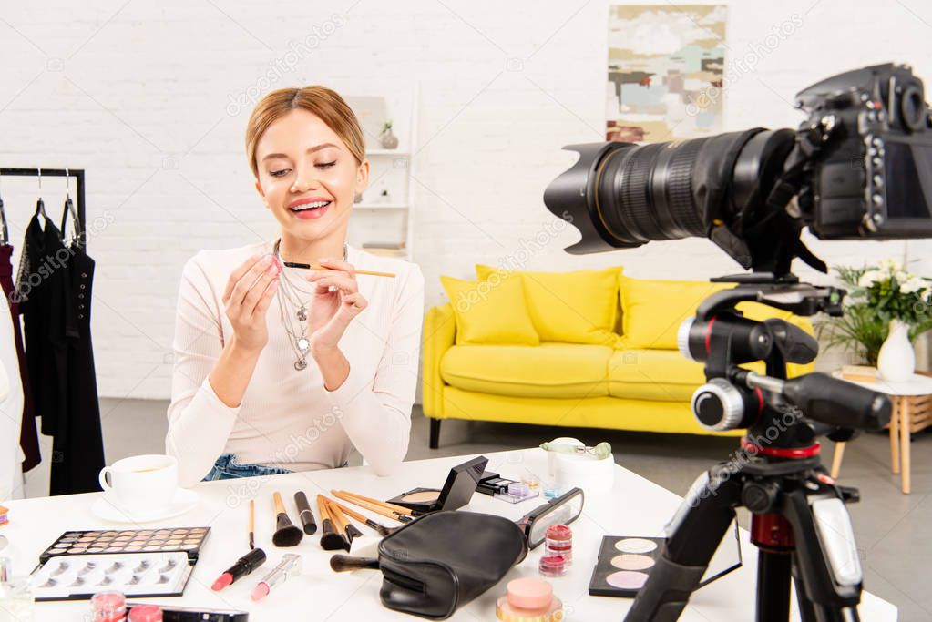 smiling beauty blogger holding lip gloss and cosmetic brush in front of video camera
