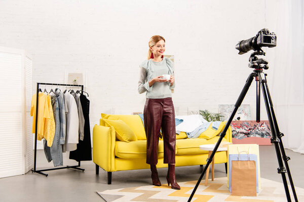 full length view of smiling blogger holding cup of coffee in front of video camera at home