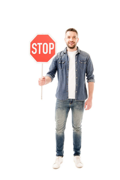 full length view of smiling bearded man man in denim shirt holding stop sign isolated on white
