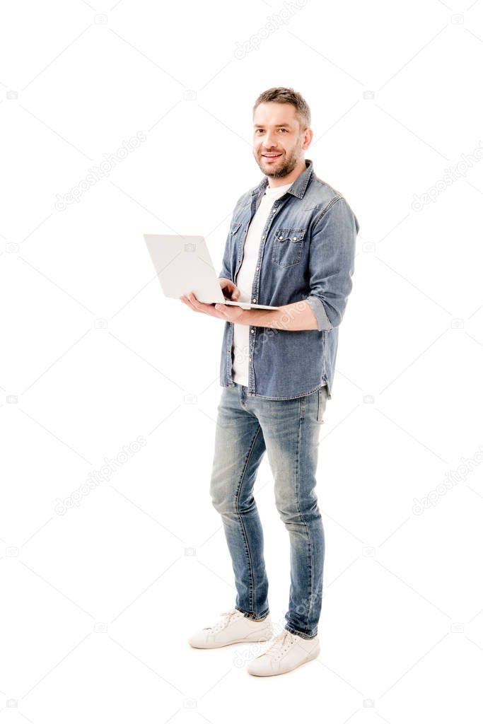 full length view of smiling man in denim shirt holding laptop isolated on white