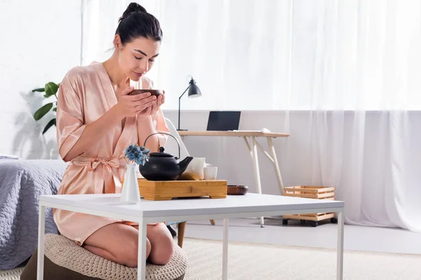 Woman in silk bathrobe with wooden bowl in hands having tea ceremony in morning at home — Stock Photo