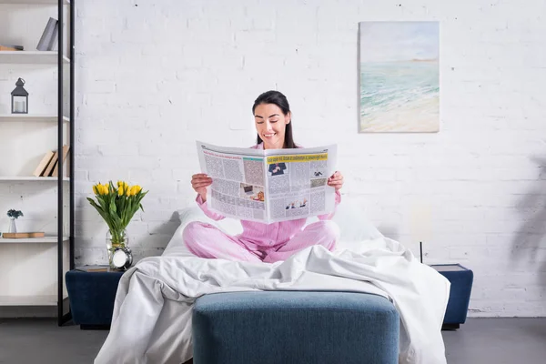 Smiling woman in pajamas reading newspaper in bed at home — Stock Photo