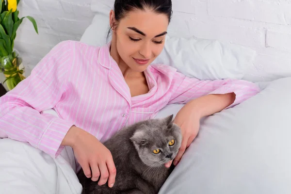 Portrait de femme en pyjama avec chat à poil court britannique reposant sur son lit à la maison — Photo de stock