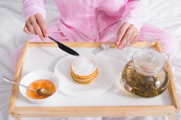 Partial view of woman in pajamas having pancakes with jam for breakfast in bed at home — Stock Photo