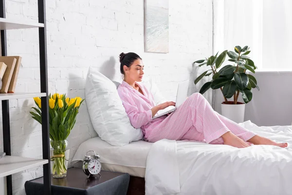 Vista lateral de la mujer en pijama usando el ordenador portátil en la cama en casa - foto de stock