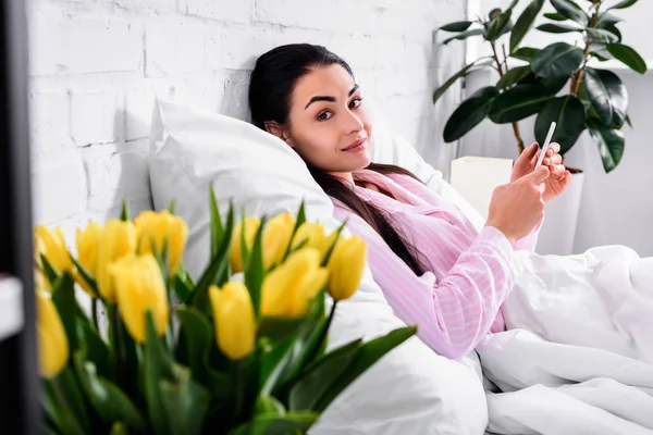 Selective focus of attractive woman in pajamas with smartphone resting in bed at home — Stock Photo