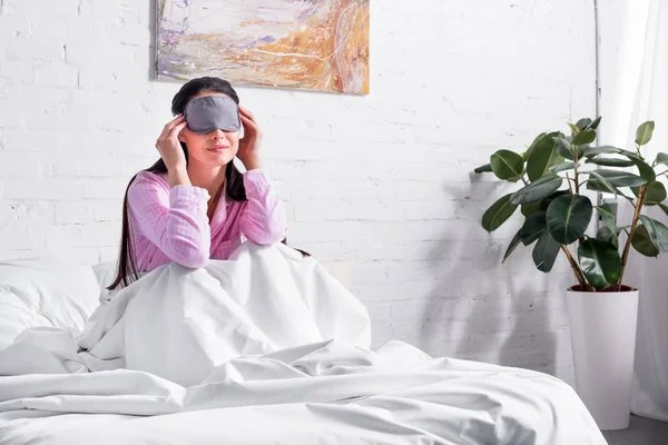 Mujer en pijama rosa y máscara de dormir sentada en la cama en casa - foto de stock