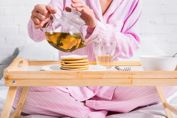 Teilbild einer Frau im rosafarbenen Pyjama, die grünen Tee in Tasse gießt, während sie zu Hause im Bett frühstückt — Stockfoto