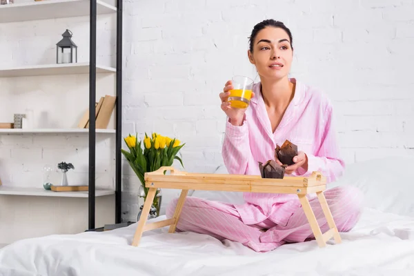 Woman in pajamas with glass of orange juice and chocolate muffins on bed in morning at home — Stock Photo