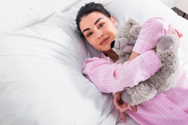 Portrait de femme en pyjama rose avec ours en peluche reposant dans son lit à la maison — Photo de stock