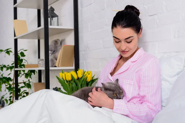 Woman in pajamas looking at britain shorthair cat on hands while resting on bed at home — Stock Photo