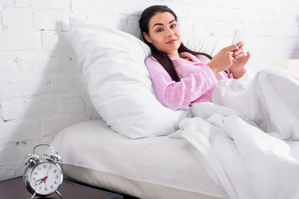 Привлекательная женщина в пижаме со смартфоном во время отдыха в постели дома — стоковое фото