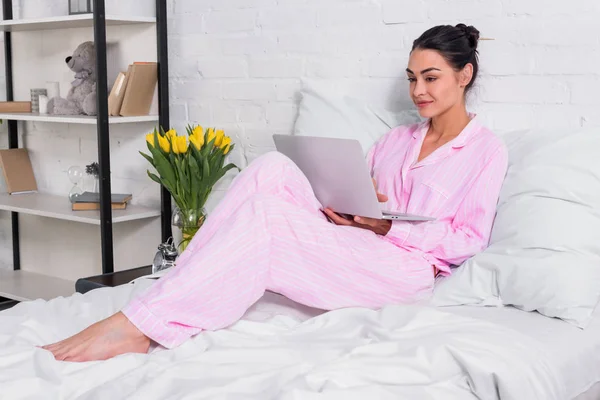 Улыбающаяся женщина в пижаме с ноутбуком в постели дома — стоковое фото
