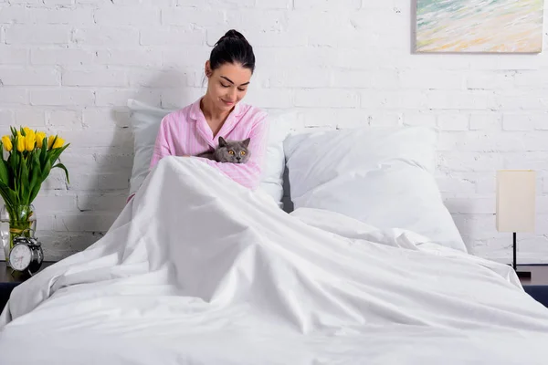 Улыбающаяся женщина гладит британскую короткошерстную кошку во время отдыха в постели дома — стоковое фото